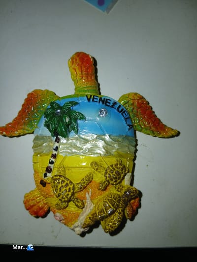 A souvenir magnet shaped like a sea turtle is displayed on a plain, light-colored surface. The turtle magnet is colorful, with a yellow and orange shell, green wings, and a blue background depicting a sky with a palm tree and the word "VENEZUELA" in black letters. The scene on the turtle's shell shows a beach with sand and several smaller sea turtles. The bottom left corner of the image has text that reads "Mar..." followed by a wave emoji. This souvenir likely represents the coastal regions of Venezuela, which includes areas like La Tagua.