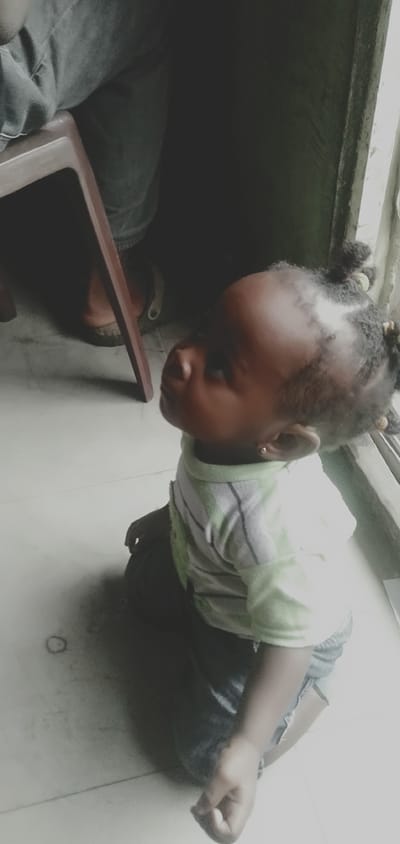 A young Black child is kneeling on a tiled floor, looking upwards and to the right. The child has dark skin, black braided hair with yellow bands, and is wearing a light green and white striped t-shirt and dark shorts. The child has a small earring in their right ear. The child is positioned in the foreground of the image.
Behind the child, a dark wooden chair is visible. To the left of the chair, the legs of an adult wearing dark trousers and light-colored sandals are partially visible, suggesting they are seated. The background is dark and indistinct, with a window frame visible on the right side of the image, suggesting the scene is indoors near an opening. The floor appears to be white or light-colored tile. The lighting suggests it might be daytime, with light coming from the window. The image was taken in Shangisha, Nigeria.