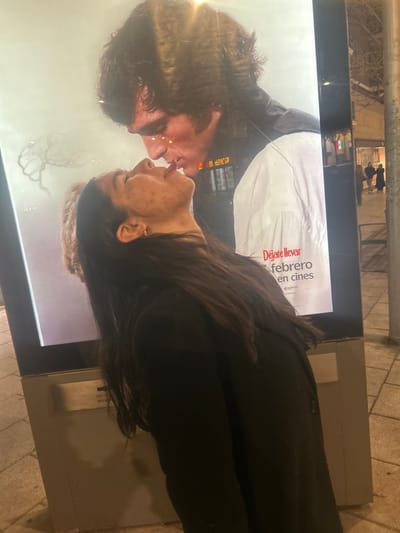 A woman stands next to a backlit poster of a couple kissing. She has long, dark hair and is wearing a black jacket. The poster's text is in Spanish, advertising a film release in February. The street scene is visible in the poster's reflection, including buildings and blurry figures. The woman's face is close to the poster's image of the kissing woman.