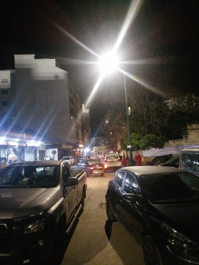 A street at night, illuminated by a bright streetlamp. Cars are parked on either side of the road. Buildings line the left side, with storefronts visible. Other vehicles are seen in the distance, along with pedestrians. The sky is dark, contrasting with the intense street light.