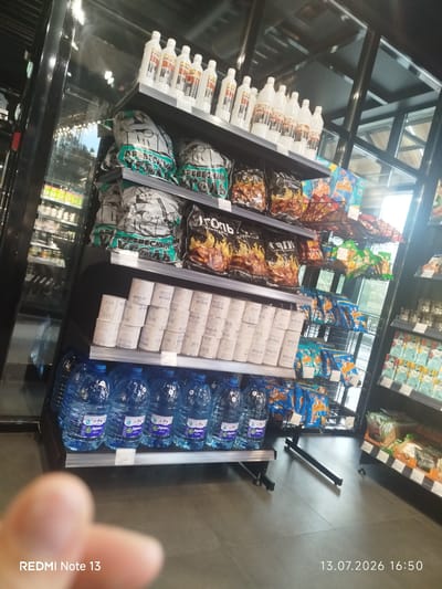 The image shows a retail environment, likely a convenience store or supermarket, with shelves stocked with various products. On the top shelf, there are several white plastic bottles, possibly cleaning supplies or beverages, arranged in rows. Below this, there are large bags of charcoal, some with green and white packaging featuring text in Cyrillic script. Further down, there are stacks of white cylindrical items, appearing to be toilet paper or paper towels. The lower shelves are filled with large blue plastic bottles of water, and a variety of snack bags and packaged goods, including chips and other food items. The overall lighting suggests an indoor commercial setting. The bottom of the image displays text indicating "REDMI Note 13" and a date and time stamp: "13.07.2026 16:50". No people are clearly visible in the image. The environment appears to be well-lit and organized, typical of a retail display. The products suggest a focus on household items, beverages, and snacks.