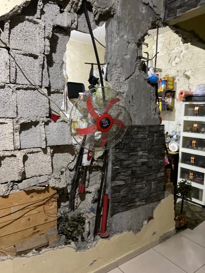 The image depicts the interior of a building in Khan Yunis, Palestinian Territories, severely damaged, possibly due to conflict or structural failure. The walls are partially collapsed, revealing the brickwork and reinforcing bars. Debris is scattered throughout. An electric fan is suspended amidst the wreckage. In the background, intact sections of the building show furniture, appliances, and household items. The scene suggests a destroyed or uninhabitable space.