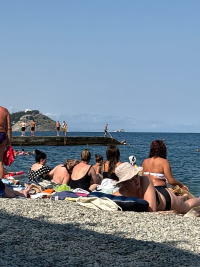 In the city of Partenit, Ukraine, a crowded pebble beach is depicted on a clear, sunny day. The foreground is dominated by a dense scattering of smooth, grey and white pebbles, casting stark shadows from an unseen source. A diverse group of people are spread across the beach, many seated or lying on towels and inflatable mats.
In the mid-ground, several individuals are engaged in various beach activities. Some are wading in the calm, blue sea, while others are sunbathing. A concrete pier extends into the water, with several men standing on it, facing the sea. One man is captured in mid-action, appearing to be preparing to jump into the water. The sea stretches out to the horizon, meeting a pale blue sky. In the distance, a large, rocky hill with some structures on its summit is visible, a prominent geographical feature of the Partenit coastline. The overall atmosphere is one of a typical summer day at a popular seaside resort.