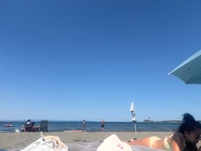 The image depicts a sunny day at Atiya Beach, Bulgaria. In the foreground, a person is lying on a towel, with a beach umbrella partially visible. The sand is light brown. In the mid-ground, several people are enjoying the beach and the sea. Two young boys are playing near the water, with one standing next to an orange surfboard. Further down the beach, a person is seated in a beach chair, and another person is standing nearby. The sea is calm and blue, with a few distant boats and individuals swimming or engaged in water activities. The horizon shows a hazy landmass. The sky is a clear, bright blue, dominating the upper portion of the image.