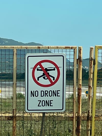 A "NO DRONE ZONE" sign is prominently displayed on a weathered metal fence. The sign features a red circle with a diagonal line through a silhouette of a drone, indicating a prohibition on drone operations. Below the graphic, the words "NO DRONE ZONE" are printed in black capital letters. The fence itself appears to be constructed of rusty metal bars and mesh, suggesting an outdoor, possibly industrial or restricted area. In the background, there are hints of distant hills or mountains under a clear blue sky, and some scattered structures. The scene suggests a location where drone use is not permitted, possibly due to proximity to an airport, sensitive infrastructure, or other safety concerns. The precise location in Mrčevac, Montenegro cannot be determined from the image alone.