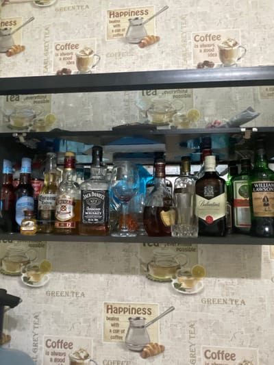 The image shows a liquor cabinet stocked with various alcoholic beverages. Brands visible include William Lawson's, Jagermeister, Ballantine's, Jack Daniel's, Chivas Regal, and Corona Extra. The cabinet is set against a backdrop of patterned wallpaper featuring tea and coffee motifs. The image suggests an indoor setting, possibly a home or bar. There's no indication of location in Sarapul, Russia, or any people or actions depicted.