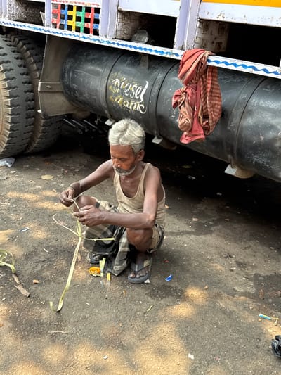 An older man with white hair and a beard is crouched beside a large vehicle, possibly a truck or lorry, on a dusty, uneven ground. He is wearing a light-colored tank top and a plaid lungi, with dark sandals on his feet. His skin appears tanned or darkened from sun exposure, and his expression is focused on the task at hand.

The man is actively manipulating a long, thin, light-yellow strip of material, appearing to be either plastic strapping or a sturdy fabric band, which has "KATHUAA COTTON" (partially visible and inverted) printed on it. He holds it with both hands, suggesting he is tying, fastening, or preparing it for use.

The vehicle next to him features large black cylindrical tanks underneath, one of which clearly has "Diesel Tank" written in yellow script. Part of the vehicle's metal framework, painted in white with a blue wavy pattern and colorful gridded sections (red, green, blue, yellow), is also visible. A crumpled reddish-brown and white striped cloth is tied or draped over a metal bar of the vehicle's frame.

The setting appears to be an outdoor, industrial or roadside area, likely a mechanic's or transport hub, common in places like Kolkata, India. The ground is dry, a mix of dirt and scattered debris including small pieces of plastic, paper, and wood. Bright, direct sunlight illuminates the scene, indicating daytime, possibly mid-day or afternoon. The overall impression is one of a laborer engrossed in his work.
