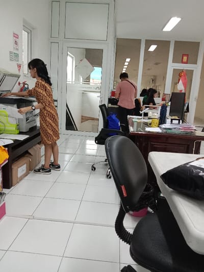 The image shows an office interior with a woman in a brown and gold dress operating a printer. The printer is on a desk with a white tiled floor. There is another desk in the background with a man in a pink shirt sitting at it. A blue bag is on the floor next to a black office chair in front of the desk. The office is well-lit and appears to be in a professional setting. The image appears to be taken from a low angle, close to the printer and the woman. The location, based on the coordinates provided, is in Kecamatan Ngaliyan, Semarang, Indonesia.