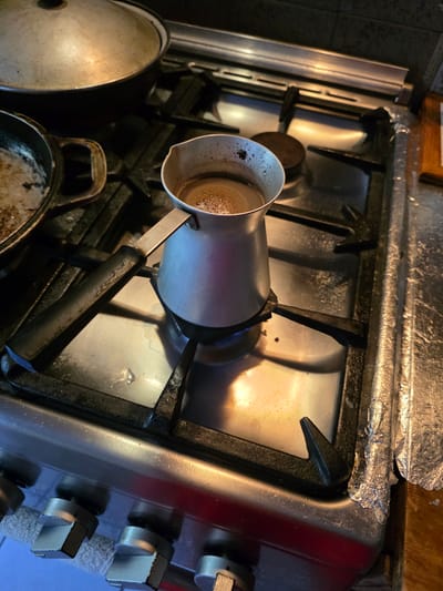 This media file depicts a close-up, overhead view of a gas stove in a kitchen setting. The primary subject is a moka pot, a stovetop coffee maker, positioned on one of the burners. The pot is made of metal and appears to contain some liquid with a foamy surface, suggesting that coffee is either being brewed or has recently been brewed.
Other objects visible include other pots and pans on the stove, some of which appear to be cast iron, and a metal lid. The stove itself is a standard gas model with metal grates and control knobs. The surface of the stove shows signs of use, with some splatters and a generally unpolished appearance. There's also a piece of aluminum foil haphazardly placed on the backsplash above the burners, possibly to catch grease or protect the surface.
The lighting in the scene is warm and somewhat dim, with highlights reflecting off the metal surfaces of the stove and cookware. This suggests it might be indoors during the evening or in a room with limited natural light. The overall impression is of a functional, lived-in kitchen. There are no people visible in the image.
The location context provided is Tuapse, Russia, but there are no specific visual cues within the image that definitively confirm this location. The scene is consistent with a kitchen found in many parts of the world.
There is no visible text in the image.