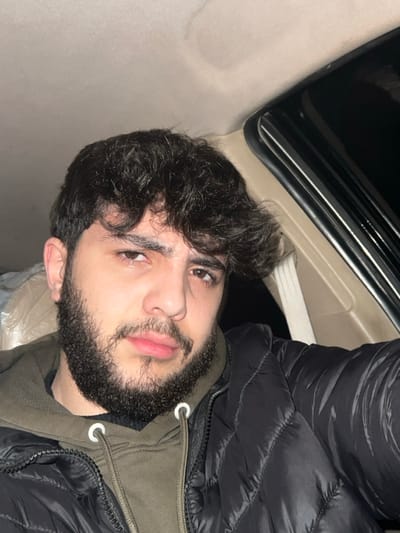 A young man with dark, curly hair and a full beard gazes at the camera. He is wearing a black puffer jacket over a green shirt. The setting appears to be inside a car, with part of the car's interior visible in the background. The lighting is somewhat dim, casting shadows on the scene. The man's expression is neutral, with a slight hint of a smile.