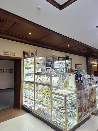 The image shows the interior of a gift shop in Peterhof, Russia. The shop features numerous glass display cases filled with a wide variety of souvenirs and gift items. The items include what appear to be nesting dolls (Matryoshka dolls), jewelry, trinkets, and other decorative objects.

The display cases are brightly lit, illuminating the merchandise within. Some of the items on display are small paintings, possibly of Russian landscapes or historical scenes, and small decorative eggs. In the background, a framed picture hangs on the wall, and a doorway is visible.

There are some signs in Russian. One sign near the doorway reads "МАГАЗИН" (MAGAZIN - Shop), "БАНКОМАТ" (BANKOMAT - ATM), and "SHOP". Another sign is partially visible above a display case and seems to include Cyrillic text.

Two small figurines, resembling angels or cherubs, are placed on top of one of the display cases. An umbrella with a patterned design is also visible in one of the lower display cases. The overall impression is of a well-stocked souvenir shop catering to tourists. The lighting suggests it could be daytime, or the shop is well-lit internally. The decor appears to be traditional with wooden paneling on the walls.