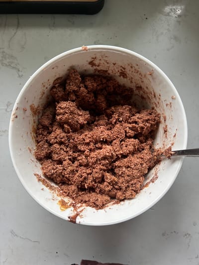 In Chang Phueak, Thailand, a white bowl contains a mixture of what appears to be chocolate and coconut. The mixture is clumpy and somewhat dry, with individual strands of coconut visible. The bowl is placed on a light-colored countertop with a subtle marble-like pattern. A metal spoon rests on the right side of the bowl, partially submerged in the mixture, suggesting it was used for mixing or scooping. The lighting appears to be natural, coming from above and to the left, casting soft shadows. There are no people visible in the image. The scene is a close-up, overhead shot focused on the contents of the bowl.