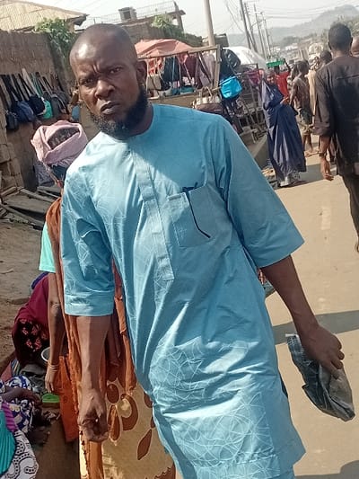 A bearded man wearing a blue, patterned long-sleeved shirt stands in an outdoor setting. Behind him, a crowded market scene unfolds with numerous stalls and people milling about. Bags are displayed prominently. In the foreground, a patterned mat lies on the ground near seated individuals. The man is holding a cloth item in his hand.
