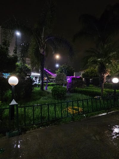 The image depicts a public park at night, characterized by wet surfaces indicative of recent rain, located in the Jardin de Florence, Ville Nouvelle, Fez, Morocco. The foreground consists of a dark, wet asphalt path reflecting light. A dark green ornamental metal fence separates this path from a grassy garden area.
Within the fenced garden, lush green grass and dark shrubs are visible. Two white, spherical streetlights on short black posts illuminate parts of the midground from within the garden, one on the left and one on the right. A coiled yellow garden hose is lying on the grass behind the fence, positioned towards the right side of the frame.
The background features several tall palm trees and dense foliage. Some of these trees or structures within the park are illuminated with distinctive purple decorative lighting. Additional streetlights, including a bright white one and a warmer yellowish one, are visible against the dark sky. Faintly illuminated buildings, including a multi-story structure on the far left with a vertical strip of white light, are visible in the distant background. No individuals are present in the scene.