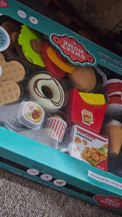 The image shows a toy set of fast food items in a teal box with the words "SWEET HOME" and "Mi primer set de comida rapida" written on it. The set includes a burger, fries, popcorn chicken, a donut, soda, a condiment container, an ice cream cone, and waffles. The box also has symbols for imagination and social skills, with a "+3" symbol indicating the recommended age. The box is placed on a carpeted surface. There are no people in the image, and there is no information related to the location of San Bernardino Tlaxcalancingo, Mexico.