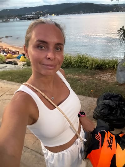 This is a vertical, low-angle, close-up selfie of a woman with short blonde hair and blue eyes. She is wearing a white tank top and white bottoms. Around her neck, she wears a delicate peace sign necklace. On her left wrist, she has a pink smartwatch. To her right, a bright orange and black JPX helmet is visible, alongside two large black garbage bags. The background shows a body of water, possibly the sea, with a distant coastline featuring lush green hills and lights from a town or village. The sky appears overcast, suggesting it might be late afternoon or evening. The ground beneath her is paved with cobblestones. The setting suggests an outdoor location in a tropical environment, potentially near a beach or waterfront area in Baan Plai Laem, Thailand. The woman's expression is neutral, and the overall mood of the image is casual.