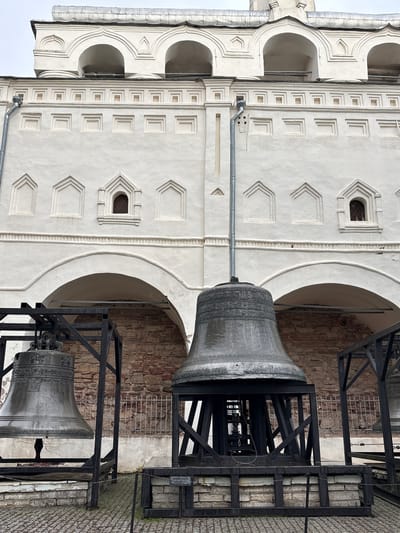 The image shows a wide, low-angle shot of the bell tower of the Saint Sophia Cathedral in Veliky Novgorod, Russia. The cathedral's white stone facade, adorned with arched windows and decorative carvings, dominates the upper portion of the frame. In the foreground, three large bronze bells are displayed on black metal stands. The bells are intricately decorated with inscriptions and figures. The ground in front of the bells is paved with cobblestones, and a low metal fence encloses the area. The overall atmosphere is one of historical grandeur and solemnity.