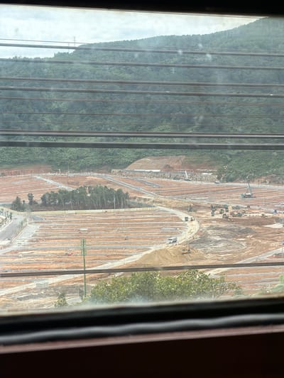 The image depicts a large-scale construction site in Hải Vân Ward, Vietnam. The terrain is mountainous and covered in lush green vegetation in the background. In the foreground, the land has been cleared and flattened, with numerous rows of demarcated plots, suggesting the development of a housing or commercial area. Several construction vehicles, including trucks, excavators, and cranes, are actively working on the site. Roads and pathways are being laid out throughout the development. A communication tower is visible in the mid-ground. The scene is viewed through horizontal blinds, which create a striped effect across the image.