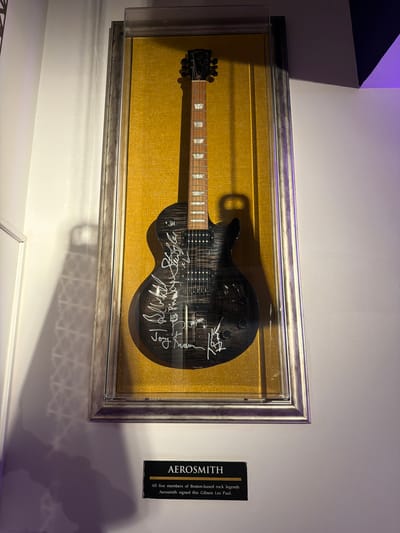 A dark-colored, signed Gibson Les Paul electric guitar, featuring a flamed top and equipped with chrome hardware, is vertically displayed within a rectangular glass-fronted case. The guitar's body is covered with multiple white signatures attributed to the band Aerosmith. The instrument is set against a textured, gold-colored fabric background inside a silver-toned frame. Directly below the display case, a black plaque with gold lettering identifies the item, stating "AEROSMITH" and noting that "All five members of Boston-based rock legends Aerosmith signed this Gibson Les Paul." The display is mounted on a plain white wall within an interior space, characterized by ambient lighting that casts subtle shadows and exhibits a purple tint on the right side. This scene is located at the Hard Rock Cafe Porto, situated in the city of Porto, Portugal. No individuals are present in the frame.