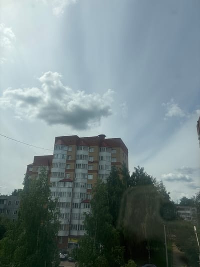 This is a vertical, eye-level shot of an apartment building in Kirov, Russia, on a cloudy day. The building is made of brick and has several stories, with numerous windows and balconies. Trees with green leaves surround the building, obscuring some of its lower sections. The sky is overcast with scattered clouds. There is no visible activity or people. The overall mood is neutral. The text "ТЕНТО" is visible on a sign on the building.