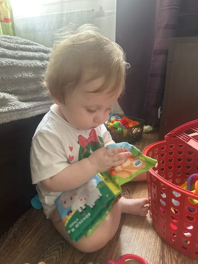 A blonde toddler, wearing a white t-shirt with a red bow graphic and two small red hearts, is sitting on a wooden floor, engrossed in a colorful soft book. The book features illustrations of sheep and other characters. To the right of the toddler is a bright red plastic basket filled with various toys, including a ring toy and other colorful objects. In the background, to the left, a gray, fuzzy blanket is draped over furniture, and a window with a radiator is visible. To the right, a dark wooden dresser or cabinet stands beside a purple curtain. The scene appears to be indoors, likely in a child's play area. The image does not provide any specific geographical markers to indicate the location within Sayanogorsk, Russia.