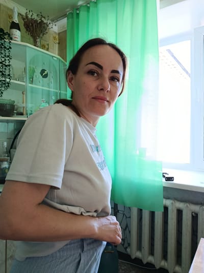 The image shows a woman in a casual setting, likely a home environment. She is positioned in the center of the frame, facing slightly towards the viewer. She appears to be wearing a light-colored t-shirt and light blue striped pants. Her hair is pulled back, and she has dark eyebrows and light-colored eyes. Her skin tone is fair.
In the background, to the left, there are kitchen cabinets with glass doors. A bottle of champagne and a vase of dried flowers are visible on top of the cabinets. To the right, there is a bright window with green curtains partially drawn. A white radiator is visible below the window. The overall lighting suggests an indoor environment during the day. The scene does not contain any specific indicators that would allow for pinpointing the location within the city of Kokshetau, Kazakhstan.