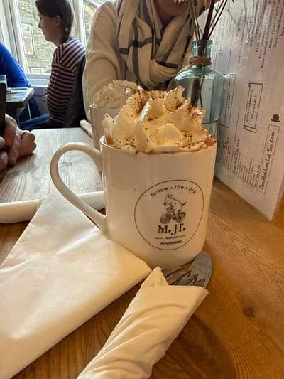 The image displays an interior scene from Mr H's Tearoom in Ambleside, United Kingdom. The central focus is a white ceramic mug, branded with "Mr H's Tearoom - Ambleside" and a "follow the pig" logo featuring a pig on a bicycle. The mug contains a dark beverage generously topped with whipped cream, dusted with cinnamon or cocoa powder. A second, similar mug with whipped cream is partially visible behind it. These mugs rest on a light-colored wooden table. A folded white paper napkin lies beneath the prominent mug, and a rolled napkin containing cutlery is positioned to its right. A light blue-green glass bottle, wrapped with twine and holding bare twigs, acts as a centerpiece. Behind it, a white menu sheet or stand displays various items and prices, including "ICED COFFEE'S AVAILABLE" and "Breakfast loose leaf Tea £3.25". In the mid-ground, a female individual with dark hair, wearing a horizontally striped shirt, is seated at a table by a window, looking left in profile towards stone buildings outside. Another person is partially visible opposite the main subject, wearing a light-colored top and a striped scarf. A person's hand and forearm, wearing a blue sleeve, are visible in the left foreground of the table.