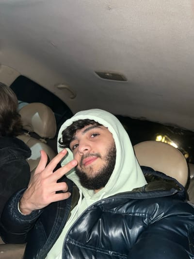 A young man wearing a light green hoodie and a dark puffer jacket is sitting in what appears to be the backseat of a car. He is looking directly at the camera with a slightly amused expression, and his left hand is raised, showing three fingers. The interior of the car is visible, including the ceiling, seats, and a grab handle. The lighting suggests it might be nighttime.