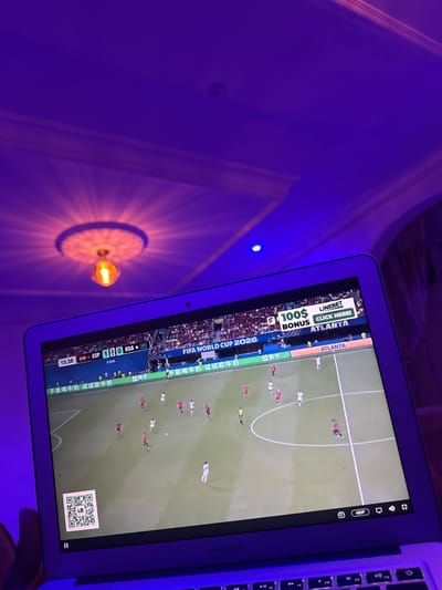The image shows a close-up of a laptop screen displaying a soccer match. The screen is illuminated by purple ambient lighting, casting a glow on the laptop's keyboard and the surrounding ceiling. On the screen, a soccer game is in progress, with players in red and white uniforms on a green field. A scoreboard at the top of the screen indicates "ESP 1 0 KSA", suggesting a match between Spain and Saudi Arabia. Above the game, an advertisement for "LINEBET" with "$100 BONUS" and "CLICK HERE!" is visible, along with the text "ATLANTA". In the bottom left corner of the screen, a QR code is displayed. The laptop appears to be a MacBook Air, as indicated by the branding on the keyboard. The overall scene suggests a person is watching a soccer match on their laptop in a room with purple mood lighting, possibly in Laminga, Nigeria.