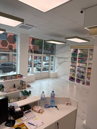This is an interior photograph of a pharmacy in Madan, Bulgaria. The pharmacy appears modern and well-lit, with white walls and shelves stocked with various medications and health products. The floor is covered in white marble-patterned tiles.

Prominently displayed on a shelf is the pharmacy's name, "Ogapharm," in elegant, dark lettering. On a counter, several items are visible, including two large bottles of water, a bag of "bebo" branded snacks, a calculator, a blue pen, and various papers and receipts. A pair of scissors is also on the counter.

Through the large glass doors and windows, the exterior of the building and street scene are visible. Cars are parked outside, and the sky suggests it is daytime. One window displays a digital thermometer showing "25°C," indicating pleasant weather. A security camera is mounted on the ceiling. The overall impression is that of a clean, organized, and functional pharmacy ready to serve customers.