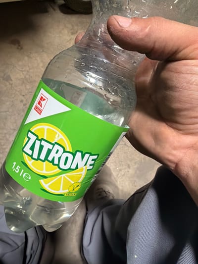 The image shows a close-up view of a hand holding a plastic bottle of "Zitron" (lemon) flavored drink. The bottle has a green label with a depiction of lemons and the text "ZITRONE" and "1,5 le". A red and white logo with "CLASSIC" is also visible. The hand is holding the bottle near the top. The background is a neutral, slightly textured surface. The image appears to be taken in an indoor setting, possibly a room or a storage area, with a concrete floor visible in the lower portion of the frame. There is no indication of the specific location in Osnabrück, Germany.