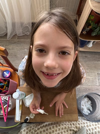 This is a candid, overhead shot of a young girl sitting at a wooden table, engaged in what appears to be a nail art activity. The girl is the primary subject, with her face filling a significant portion of the frame, looking directly at the camera with a bright, open-mouthed smile and a mischievous glint in her brown eyes. Her long, brown hair frames her face, with some strands falling forward.

On the table in front of her are various items associated with nail care and art. These include a white bottle with a brush applicator, a small glass bottle, a dark-colored nail polish bottle, a white tissue, a bottle with a green cap, and a nail file. There is also a pink container filled with colorful markers or colored pencils. A small blue jar with white and pink text is visible, which appears to be a cosmetic product, possibly Nivea.

The background shows a wooden floor and a portion of a patterned fabric, possibly a blanket or bedding. To the upper right, a grey mesh trash can is filled with crumpled white tissues. In the upper left corner, a shelf with a potted plant is visible. The lighting suggests it is daytime. The overall mood is cheerful and playful, captured during a moment of personal grooming and creative expression. The visible text on the blue jar is "NIVEA" and "CREME". The location is Zassovskaya, Russia.