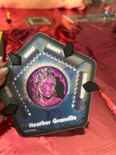 A hexagonal game piece for a board game features a portrait of a woman with pink hair and a pearl necklace against a dark blue background. The woman's name, "Heather Granville," is printed in white text at the bottom of the piece. The piece also displays various stats and information, including "MIGHT," "SANITY," "SPEED," "KNOWLEDGE," "Age: 18," "Height: 5'2"," "Weight: 120 lbs," and "Hobbies: Television, Shopping." The text "© 2021 Hasbro." is also visible. The game piece is held in a person's hand, with fingers visible on the left side of the image. The background consists of a shiny, crinkled red surface, possibly a tablecloth or wrapping paper. The overall scene appears to be indoors, with some blurry elements in the background that suggest other game pieces or objects on a table. There is no information in the image to suggest a location in Puebla, Mexico.