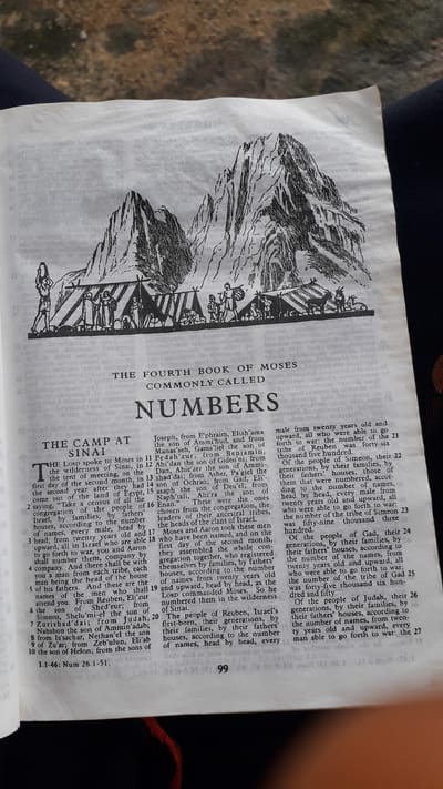 The image shows a page from a book, likely a Bible, with a black and white illustration at the top depicting a mountainous landscape with tents and figures of people and animals. Below the illustration, the text reads "THE FOURTH BOOK OF MOSES COMMONLY CALLED NUMBERS". The main body of the page contains text arranged in columns, detailing names, genealogies, and census information. The page is partially opened, with a red thread visible at the bottom right, suggesting it is a bound book. The surrounding area is blurred and appears to be a floor or surface. There is no information within the image that indicates a location in Nkpor, Nigeria.