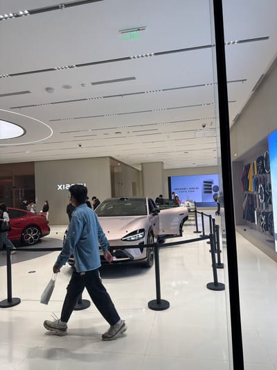 The image shows a person wearing a light blue denim jacket, black pants, and gray sneakers walking in what appears to be an automotive showroom in the Jiangning District, China. Several cars are on display, including a pink car with a door ajar and a red car. The person holds a light-colored bag. Barriers with black poles and bases are set up around the cars. A large display screen is in the background, along with the word "Xiaomi" on a wall. The showroom has a modern design with a bright, white ceiling featuring recessed lighting. The image is taken through a window or glass panel.