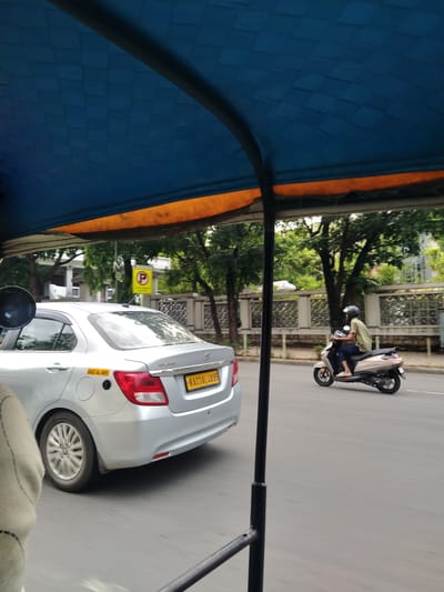 Captured from within an auto-rickshaw, the scene shows a silver Maruti Suzuki Swift Dzire taxi (registration number KA03L3955) and a scooter with a rider wearing a helmet, both navigating a Bengaluru street. The surrounding environment features trees and buildings, indicative of an urban setting in Bengaluru, India. A yellow traffic sign is also visible near the car.