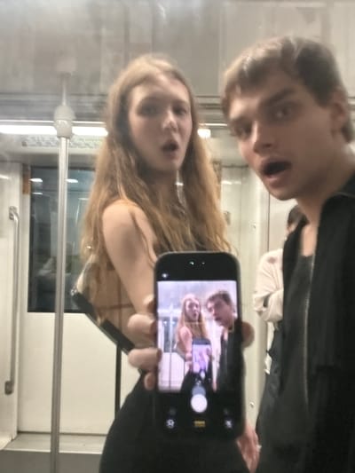 This media file is a selfie taken by two young people, likely teenagers or young adults, in a public transportation setting. The image is oriented vertically with the subjects appearing upside down. In the foreground, a young woman with long, blonde hair is visible. She is looking towards the camera and appears to be smiling or making a surprised expression. She is wearing a plaid-patterned top. Behind her and slightly to the left is a young man. His face is also visible, and he has a similar surprised or playful expression. He is wearing a dark jacket.
The subjects are holding a smartphone, which is capturing their reflection. The phone screen is visible and shows a smaller reflection of the two individuals, indicating they are taking a selfie. The background suggests they are inside a train or subway car, as evidenced by the windows, seating, and metal poles commonly found in such environments. The lighting appears to be artificial, consistent with an indoor setting. The overall mood seems lighthearted and playful. There are no visible text elements in the image. The image appears to have been taken during the day, given the ambient light.
