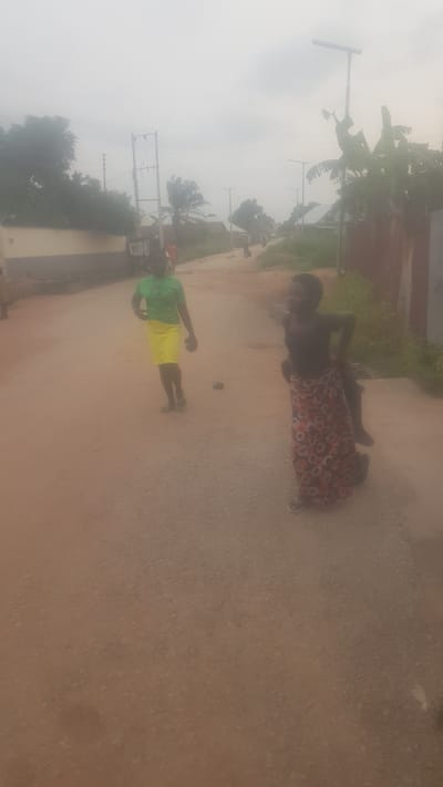 The image depicts an outdoor scene on a dirt road in Wukari, Nigeria. Two young women are the central figures. The woman on the left wears a green t-shirt and a yellow skirt, and she is looking towards the right. The woman on the right is wearing a dark top and a patterned skirt, and she is facing away from the camera, with her right arm raised. There are other individuals and a vehicle faintly visible in the background. The road is unpaved and appears to be surrounded by vegetation and buildings. The sky is overcast, suggesting cloudy weather.