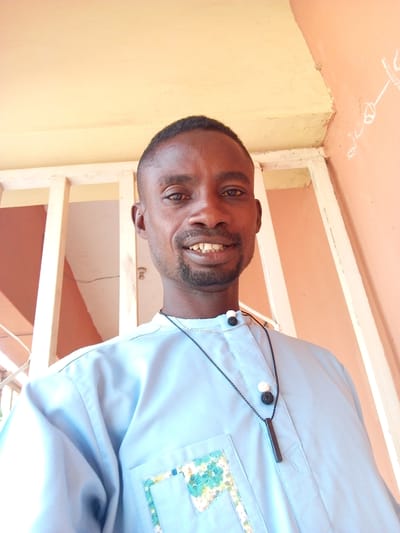 A medium shot depicts a dark-skinned man with short, dark hair and a goatee smiling at the camera. He is wearing a light blue collared shirt with a floral pattern on the pocket and a black necklace with a white and black pendant. The background features a white metal railing, a pale yellow ceiling, and a peachy-orange wall with some white markings. Given the instruction about Otokiti, Nigeria, the image is likely taken in a residential or public area within that city.