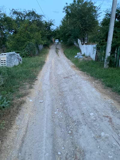 The image depicts a narrow, unpaved dirt road leading uphill through a rural area. The road surface is textured with small stones and tire tracks. To the left of the road, a stack of grey concrete blocks is visible, situated on overgrown grass. Beyond the blocks, dense green foliage and trees form a wall. To the right of the road, a weathered white wooden fence runs parallel to the path, with some sections appearing broken or dilapidated. Behind the fence, more trees and vegetation are present. In the distance, at the crest of the hill, a white van is partially visible. A grey utility pole stands on the right side of the road, with overhead wires. The sky above is a dusky blue, indicating either early morning or late evening. The overall environment suggests a secluded, semi-developed or undeveloped area, possibly on the outskirts of a village. The location is identified as Horodca, Moldova.