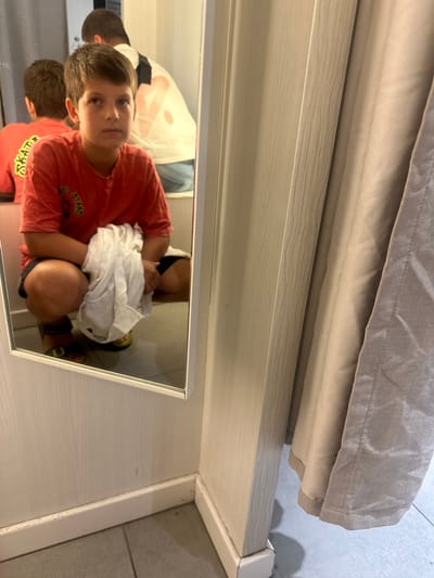 In the Polígono Industrial De Quart De Poblet, Spain, a young boy with brown hair and brown eyes is captured in a mirror's reflection. He is wearing a red t-shirt with yellow lettering that appears to say "SKATE" and black shorts. The boy is squatting down, holding a white cloth in his hands. He is in what appears to be a fitting room, with pale wood paneling and a beige curtain visible in the background. The floor is tiled in a gray, square pattern. Another person is partially visible in the mirror behind the boy, wearing a white shirt with a large graphic design. The overall lighting is moderate, and the image has a slightly grainy quality.