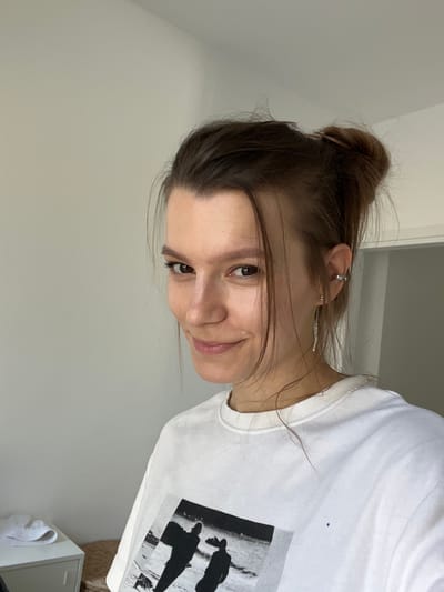 The image is a close-up, head-and-shoulders portrait of a young woman with her hair pulled up in a bun. She is looking towards the viewer with a slight smile. She is wearing a white t-shirt with a black and white graphic print on the front. She has an ear cuff and a dangling earring in her left ear. The background is a plain, light-colored wall, with a hint of a doorway visible on the right. The overall lighting suggests an indoor setting, likely a room. The image does not contain any information or visual cues that would allow for identification of its location in Limassol, Cyprus.