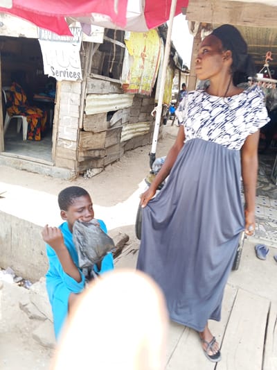 A woman wearing a grey, black, and white dress stands on a wooden surface in an outdoor setting. She is holding the sides of her dress. To her left, a child in a blue shirt sits on a concrete surface and appears to be holding something to their mouth. In the background, there is a wooden building with a sign that says "PLUS Services" and "ABU HASSAN." Underneath a red and white awning there's another building constructed of wood and corrugated metal. Other people and a vehicle are visible further back. The scene is likely located in Zango-ndaji, Nigeria, based on the prompt.