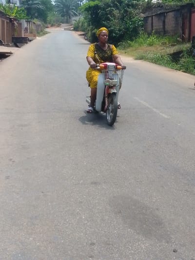 The image depicts an outdoor daytime scene on a paved road in Alor, Nigeria. An adult female is operating a red and white moped-style scooter, traveling forward on the asphalt surface. She is positioned centrally within the frame, wearing a yellow patterned short-sleeved dress, a matching yellow headwrap, and white-and-black striped slide sandals. The scooter features a silver-colored, front-mounted wire basket.
The asphalt road appears moderately worn and extends uphill, curving slightly to the left in the distance. A faded white dashed line is visible on the right side of the road from the rider's perspective.
To the left of the road, a concrete drainage ditch runs parallel to the pavement, followed by residential structures constructed with brown or orange-toned walls, some featuring visible gates. Lush green tropical vegetation, including banana plants, grows behind these structures. A small, dark-colored animal, possibly a dog or monkey, is perched on a low wall or step further down the left side of the road.
To the right of the road, a concrete boundary wall is visible, behind which dense green foliage and brush grow along an unpaved shoulder. The scene is illuminated by bright daylight, with shadows consistent with a high sun angle.