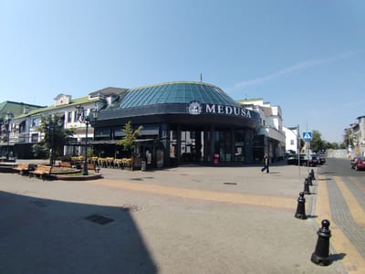 The image depicts a street scene in Brest, Belarus, featuring the "Medusa" building with a distinctive domed glass roof. The building is dark in color and features large windows and signage. In front of the building, there are several park benches and trees. The street itself is paved with cobblestones and has a yellow and grey striped pedestrian crossing. Several black bollards line the side of the pedestrian crossing. A woman is walking across the street, and a few cars are visible in the background. The sky is clear and blue. The overall impression is that of a bright, sunny day in an urban environment.