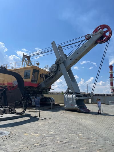 In Verkhnyaya Pyshma, Russia, at the Museum of Military Equipment "Battle Glory of the Urals," a massive, yellow and red excavator is displayed on a paved area under a blue sky with scattered white clouds. The excavator, a large industrial piece of machinery, features a prominent boom with a large red pulley at the top and a substantial grey bucket hanging from its end. The machine is complex, with numerous cables and structural elements visible. To the right of the excavator, a young boy wearing a patterned t-shirt and shorts stands near a red and white striped industrial chimney emitting a plume of white smoke, suggesting the presence of a power plant or factory in the background. The ground is paved with grey cobblestones. A small informational sign is positioned to the left of the excavator's bucket. The scene appears to be an outdoor exhibition, with a sense of scale conveyed by the immense size of the machinery compared to the human figure.