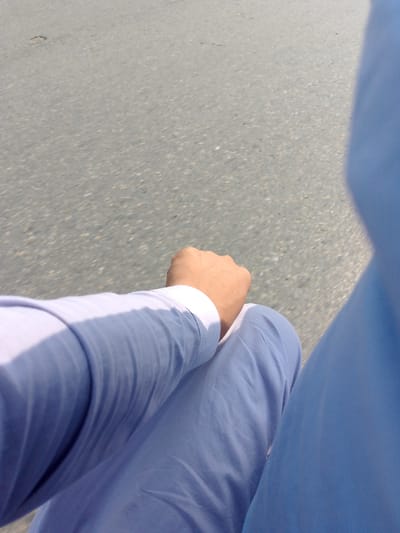The image shows a close-up view of a person's lower body and arm against a blurred asphalt background. The person is wearing light blue clothing, possibly a kurta or shalwar kameez, and their fist is clenched on their lap. The lighting suggests it is daytime, and the blurred background indicates motion, potentially from a vehicle. The setting appears to be a road in a city, consistent with Peshawar City Tehsil, Pakistan.