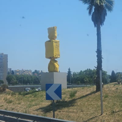 A bright yellow sculpture, resembling a wrapped candy, stands on a grey pedestal. The sculpture is composed of two stacked, distorted rectangular shapes. To the right of the sculpture, a tall palm tree with a textured trunk and drooping fronds is visible against a clear blue sky. In the background, there are buildings and dense green trees. In the foreground, a road sign with a white arrow pointing left on a blue background is planted in the ground. The scene appears to be an outdoor public space, likely near a road or highway, given the presence of the road sign and guardrail. The overall environment suggests a warm, sunny day. The location is identified as Nice, France.