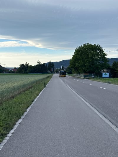 A yellow bus travels down a paved road in Lufingen, Switzerland. The road is flanked by a green field on the left and a sidewalk on the right. In the distance, trees and buildings are visible. The sky is cloudy with patches of blue.