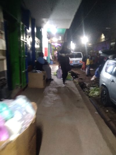 An alleyway at night with vendors selling goods from stalls. A car is parked on the edge of the alleyway. People are standing and walking, seemingly buying and selling items. There are bright lights in the alleyway, possibly streetlights or lights from the stalls. One person appears to be setting up items on the ground. The scene suggests a bustling, informal market area at nighttime.